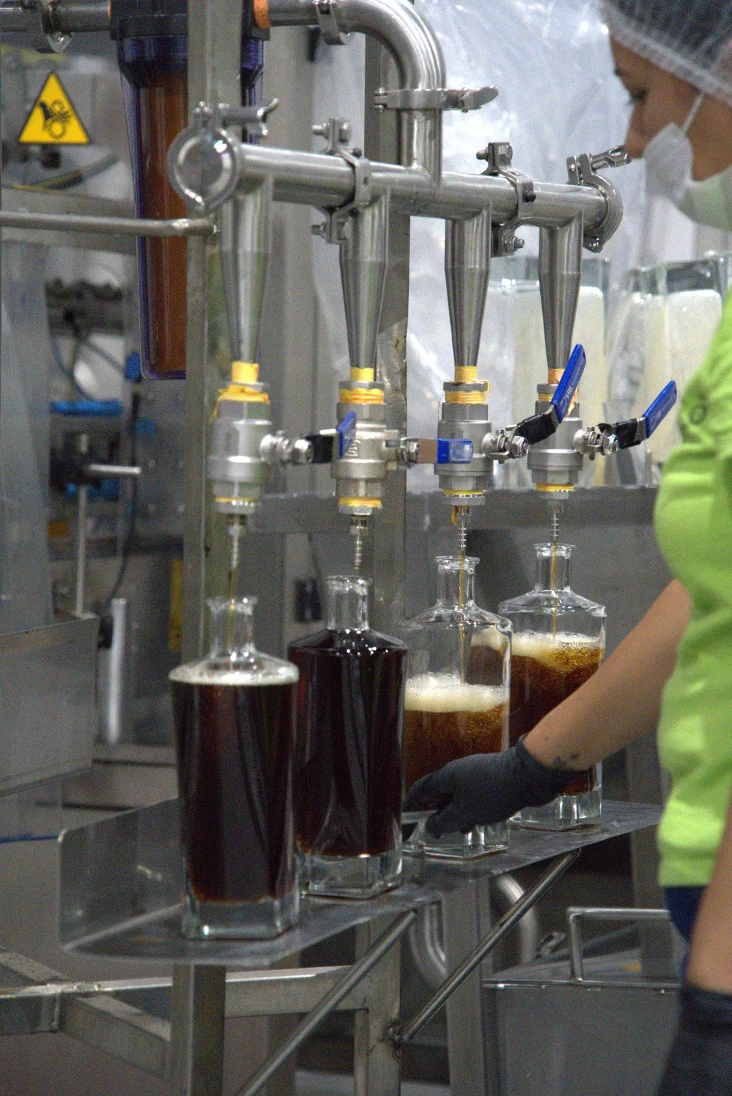 A four-head filling station running amber tequila into bottles, operated by a worker in a hairnet, mask and gloves