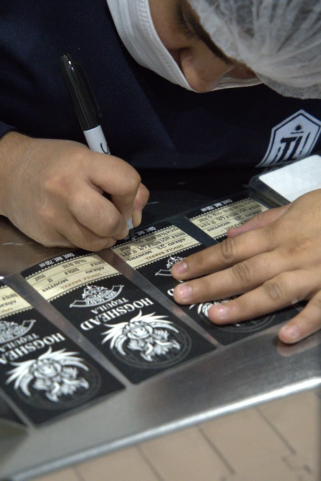 A worker writing barrel and bottle numbers by hand onto Hogshead Travesía labels laid out on a steel bench